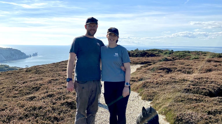 Picture of a man and a woman and a dog on a footpath near some heather with The Needles in the background